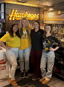 4 of the Coach's Corner staff in front of the liquor bottles, Hawkeyes lighted sign and picture